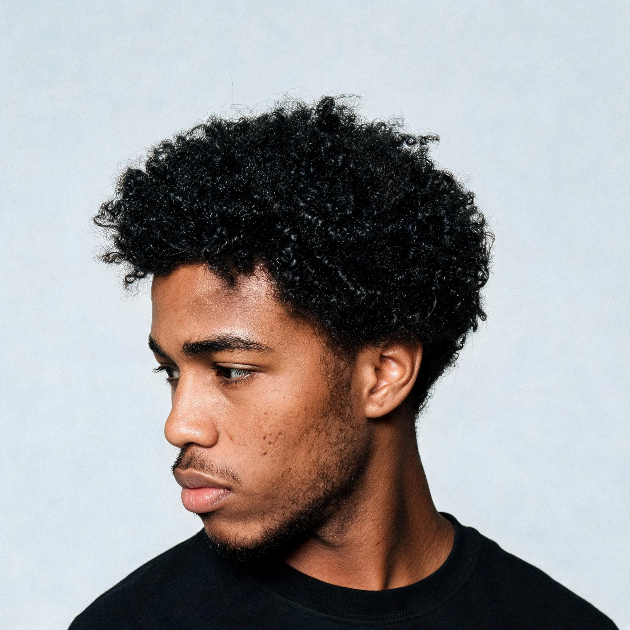 Candid editorial beauty photograph of a handsome young Black man in his early 20s, rich dark brown skin, distinctive youthful masculine features, dense natural kinky-coily black hair, medium length sculptural shape, very tight 4B/4C texture, densely packed tiny coils and zig-zag texture, visible natural strand definition, soft cloud-like volume with tactile surface texture, moisturized healthy finish without looking wet or overly glossy, natural shrinkage, irregular silhouette, realistic edges and hairline, a few stray hairs. Real skin pores, subtle blemishes and tonal variation, natural brows, faint facial hair, black crewneck t-shirt. Three-quarter profile, chin slightly lowered, eyes looking away from camera, hair silhouette fully visible. Pale cool gray seamless studio background, direct on-camera flash, authentic early-2000s editorial casting photography, medium-format film feel, subtle grain, realistic color, slightly imperfect exposure, unretouched skin, not commercial stock photography