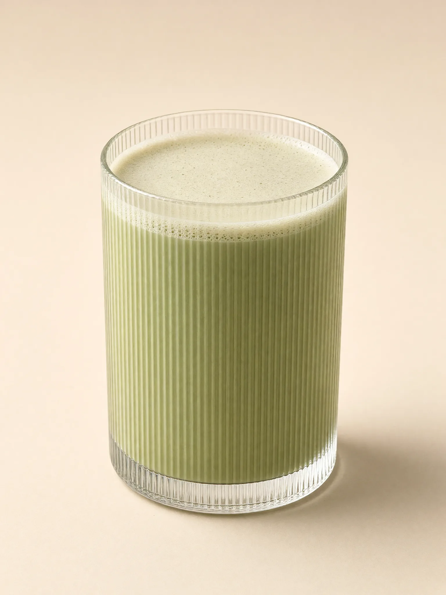 editorial photo of matcha latte in a transparent handleless cylindrical glass with a perfectly straight silhouette, flat vertical sides with no curvature, taper or expansion, covered edge-to-edge with very fine closely spaced vertical fluting from base to rim, smooth creamy latte foam visible from a gentle three-quarter elevated view toward the viewer, uniform light beige background, refined minimalist styling, soft studio reflections emphasizing the micro-faceted texture, entire glass sharply focused and uncropped. Shot on Nikon D850 with AF-S Micro NIKKOR 105mm f/2.8G IF-ED lens.