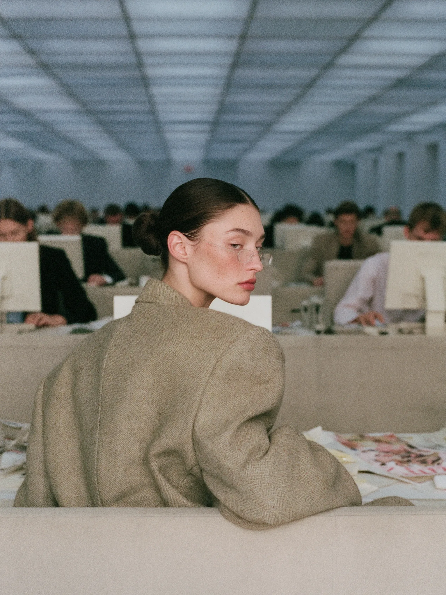 wide editorial fashion photograph of a large corporate open-plan office, rows of desks and computer monitors, several blurred coworkers in the background typing. in the middle a woman sits at her desk staring at her screen, looking away from camera in profile, calm blank expression, lips closed, slicked-back black bun, thin rectangular glasses, oversized greige wool blazer. overhead fluorescent ceiling panels, hard top light, crisp shadows, neutral white balance, clean greige and bone palette. candid documentary framing, shot on Hasselblad 50mm, f4, deep focus, ultra detailed skin texture, glossy high-end retouch, Vogue workplace editorial